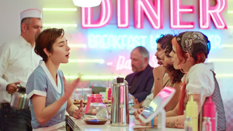Shot Of Waitress Talking To Two Non-Binary People In 1950s Styled Diner In 1950s Styled Diner