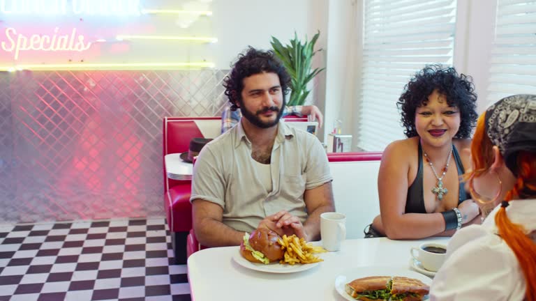 Waitress Serving Food To Two Non-Binary People And a Man In 1950s Styled Diner