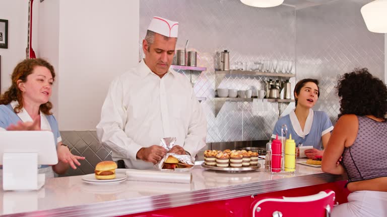 Man In White Button Down Shirt And Paper Cap Wrapping Burger In 1950s Styled Diner