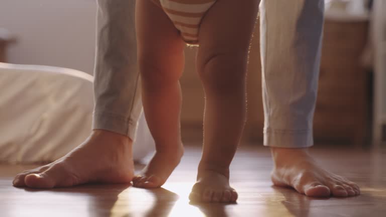 Close-up baby legs taking first steps with mother help at home. Asian little baby boy holding hands with his mother learning to walk step by step at home. Close-up baby legs taking first steps with mom.