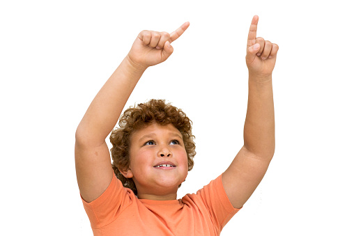 Closeup cutout of a young multiracial boy with curly red hair and an orange shirt pointing up with both fingers with excitement on a white background.