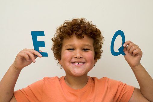 Elementary age boy with red hair and an orange t-shirt is smiling and looking at the camera while holding up the letters EQ, standing for emotional quotient or emotional intelligence.