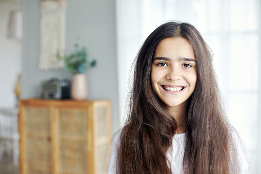 Serious beautiful preteen 12s girl pose in cozy living room, having long brunette hair smile looking at camera. Generation Alpha person portrait