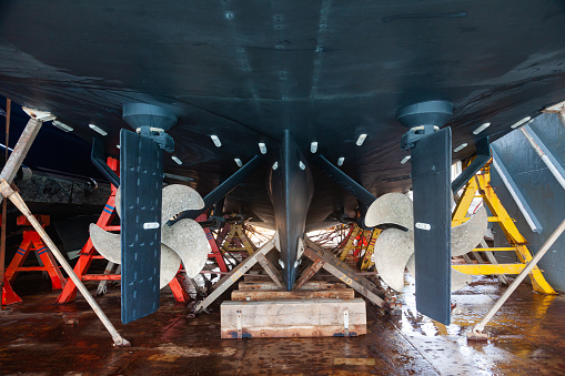 The back of a large motor yacht, standing on wooden blocks in a dry dock. Two propellers and zinc protective inserts.