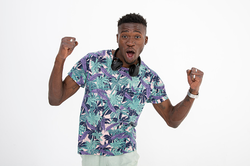 Young black man of African descent wearing patterned and colorful t-shirt, headphones, making different facial expressions. İsolated on white. Facial expression expressing Success and winning with arms raised in the air, clenching fists and opening mouth.