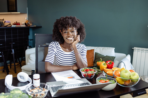 A female nutritionist is sitting at the table and looking at the camera. A laptop and plenty of fruit and vegetable are on her desk.