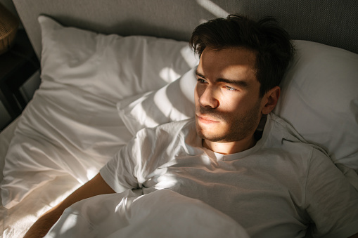 A young and handsome man lying in bed, half-darkness surrounding him, as rays of morning sunlight illuminate him through the lines on the blinds. Morning contemplating. Thinking of a day ahead of him