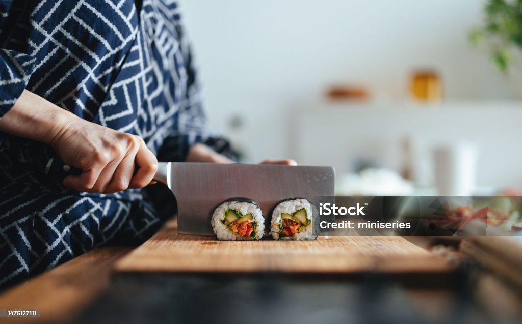 Close Up Photo Of Woman Hands Cutting Sushi Rolls Side view of unrecognizable Asian female chef is cutting sushi rolls on a wooden cutting board and preparing a healthy meal at home. Sushi Stock Photo