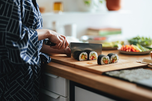 An anonymous Asian female chef is cutting sushi rolls on a wooden cutting board and preparing a healthy meal at home.