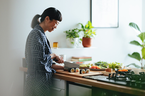 Smiling Asian female chef is cutting sushi rolls on a wooden cutting board and preparing a healthy meal at home.