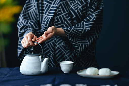 An anonymous Asian woman in kimono preparing hot tea with mochi ice cream at kitchen desk.