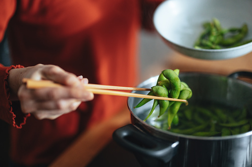 High angle view of an anonymous Asian female chef standing at the gas stove and picking up cooked edamame beans with chopsticks from the pot.