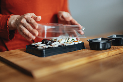 An anonymous Asian woman opening a plastic container with sushi rolls at kitchen desk.