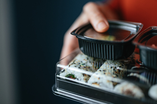 High angle view of an anonymous Asian woman holding plastic container with sushi rolls, soy sauce, wasabi and pickled ginger.