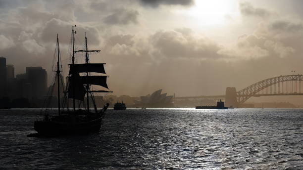 Black silhouette of a traditional barquentine or schooner barque tall ship cruising westwards the waters of Port Jackson in a very hazy afternoon-Harbour Bridge in the background. Sydney-NSW-Australia