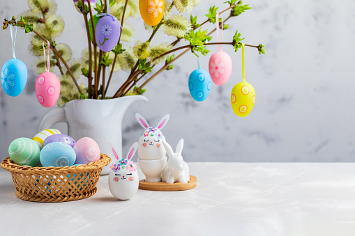 Easter composition with crafted Easter bunny, colorful eggs in a basket and flowering willow decorated with Easter eggs on a table for festive Easter celebration at home. Copy space.