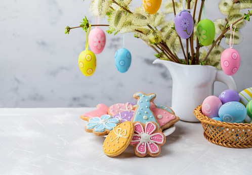 Homemade Easter gingerbread cookies shaped of Easter bunny, egg and flower, colorful eggs in a basket and flowering willow decorated with Easter eggs on a table for festive Easter celebration at home. Copy space.