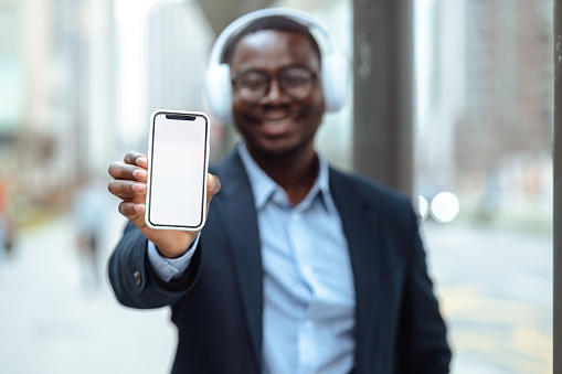 Portrait of African American good-looking man using smart phone and wireless headphones. He is holding smart phone with blank screen
