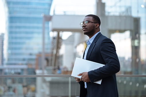 A young African-American businessman on the balcony of the business building holding a laptop and thinking