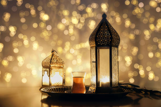 istock Festive Ramadan Kareem, Iftar dinner still life. Glowing Moroccan ornamental lanterns with glass of Turkish tea. Olive tree branch, silver tray. Blurred background, golden bokeh lights. Eid ul Fitr. 1475145981