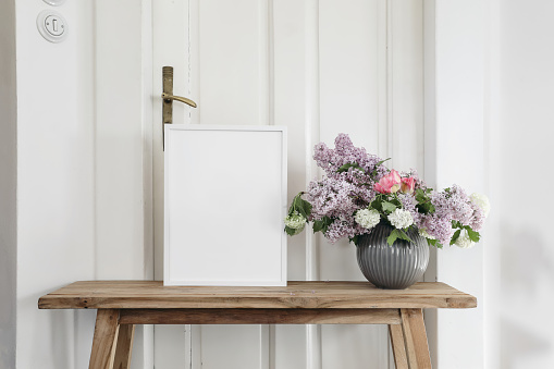 Vintage spring floral still life. Blank picture frame mockup on old wooden bench, table. Vase with bouquet of lilac, viburnum and tulips. White wall background. Empty copy space, rustic interior.