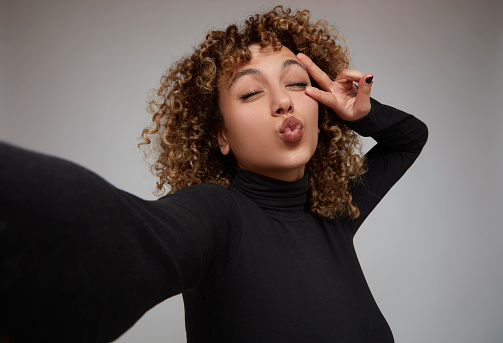 Young woman with curly hair taking selfie, making peace sign and sending kiss.