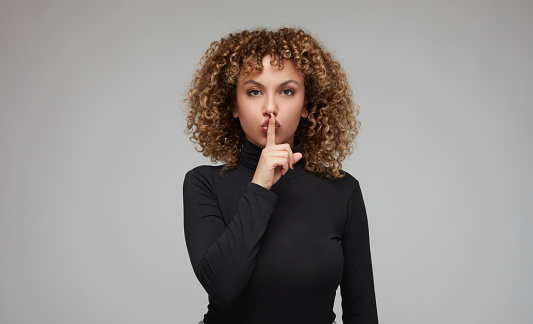 Young woman with curly hair making a hush sign with index finger on her lips.