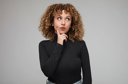 Young woman with curly hair looking away while thinking something with her hand on her chin.