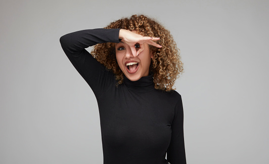 Perky fun-seeking young woman with curly hair holding her fingers round and in front of her eyes.