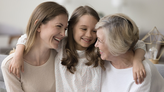 Cheerful women and kid of three different female generations meeting on home couch, hugging with love, affection, talking, chatting, laughing, enjoying family leisure, relations, bonding, having fun