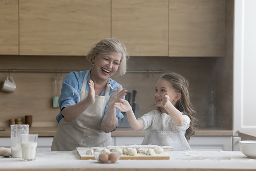 Cheerful excited grandmother and granddaughter kid having fun in home kitchen, baking, hitting floury hands over table with dough, making cloud of flour, laughing