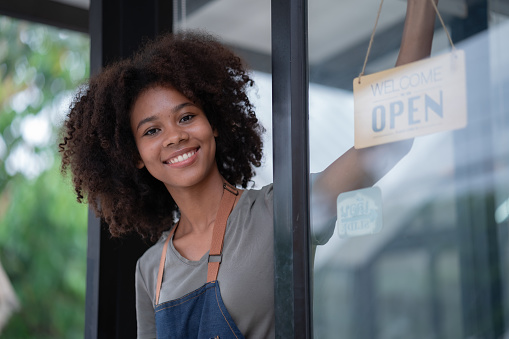 Portrait of African American businesswoman standing at the cafeteria door entrance. A cheerful young waitress in a blue apron near a glass door with an open signboard and looking at the camera.