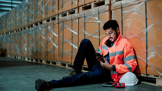 A young Asian warehouse worker sitting at a corner and browsing his phone while slacking off at work.