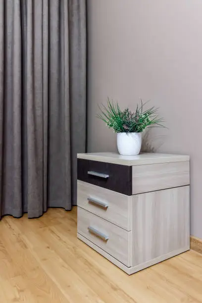 Light bedside table with a black shoe and a potted flower standing on it, against the background of the wall and curtains.
