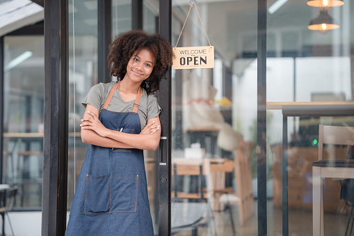 Portrait of African American businesswoman standing at the cafeteria door entrance. A cheerful young waitress in a blue apron near a glass door with an open signboard and looking at the camera.