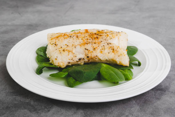 Cod fillet baked with garlic butter sauce served with fresh spinach on a white plate close-up on the kitchen table