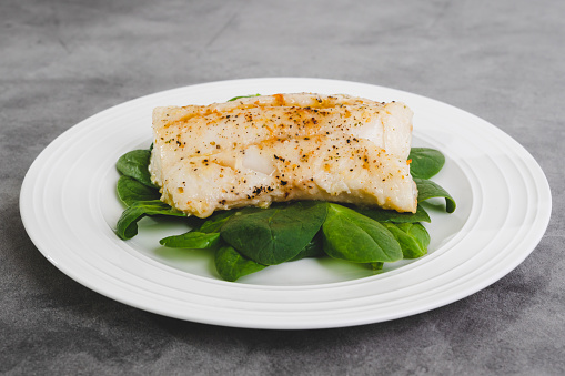 Cod fillet baked with garlic butter sauce served with fresh spinach close-up on a white plate