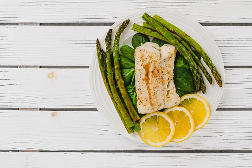 Cod fillet baked with garlic butter sauce served with fresh spinach and roasted asparagus on a white plate close-up on the wooden background, flat lay with copy space