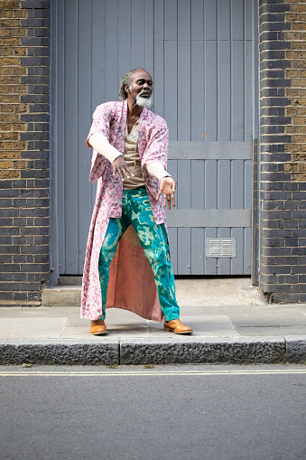 Alternative dance by a mature man in front of large grey-coloured wooden doors. He is wearing a loose-fitting pink kimono-style jacket and turquoise-coloured printed trousers.