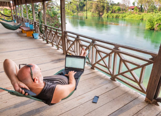 Working remotely on his computer in Si Phan Don,Southern Laos,whilst traveling the world,relaxing on a wooden guesthouse balcony overlooking the Mekong delta.Being productive in a beautiful setting.