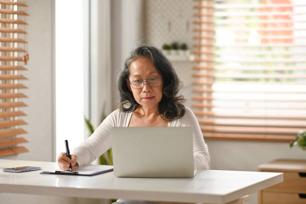 istock Focused middle aged woman watching online webinar on laptop and making notes on notebook during remote working from home. 1475215445