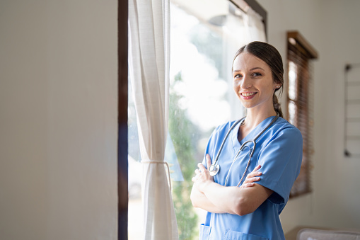 Portrait of a friendly female doctor or nurse wearing blue scrubs uniform and stethoscope, with arms crossed in hospital.