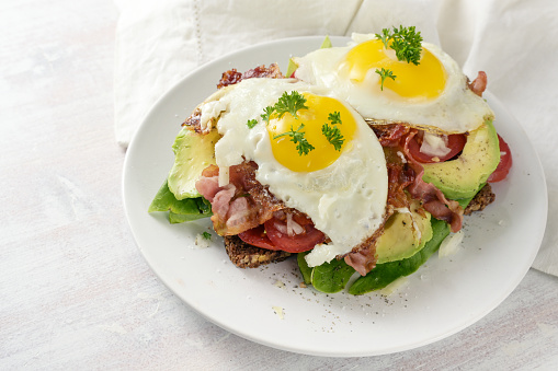 Fried egg, bacon and avocado on a dark whole meal bread with lettuce and tomato, delicious breakfast sandwich, light background, copy space, selected focus, narrow depth of field