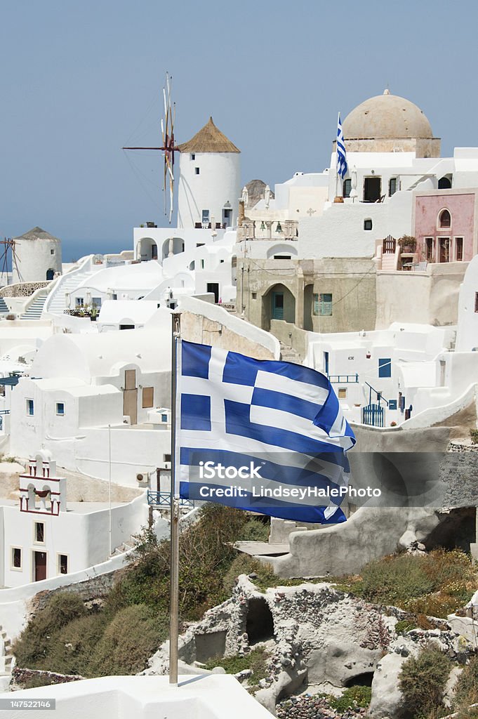 Oia Santorini A Greek flag blows in the wind on a spring day in Oia, Santorini.  The famous Greek windmills are in the background. Cyclades Islands Stock Photo
