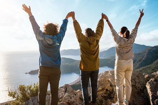 Happy female hikers taking with arms raised above the sea at sunset. Three cheerful women standing on the top of the mountain at sunset enjoying majestic view of mountains and sea. Women power concept
