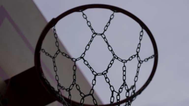 Close-up basketball ball passing hoop with metal net in slow motion at background of gray overcast sky outdoors. Score in competitive game on street.