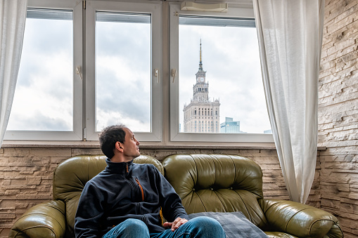 Young man sitting on couch in flat apartment looking at view of Warsaw, Poland famous Palace of Science and Culture building through window