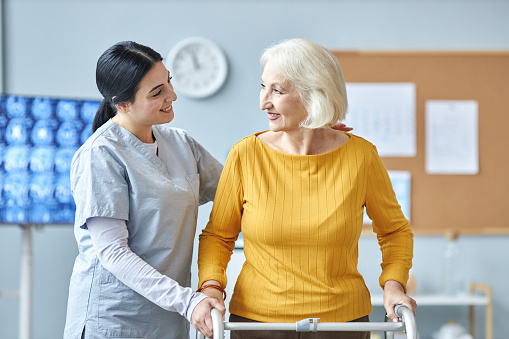 Waist up portrait of smiling young nurse helping senior woman using mobility walker in medical clinic