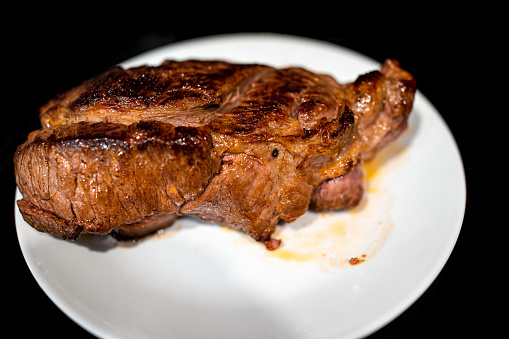 Macro closeup of cooked ribeye, strip or sirloin steak with thick cut and brown crust on white plate and background resting with juices
