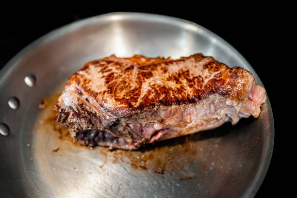 gros plan d’une poêle et d’une croûte en acier inoxydable sur une épaisse bande de new york ou un steak de viande de faux-filet cuisant sur une cuisinière avec une texture carbonisée et un fond noir - steak meat strip steak restaurant photos et images de collection